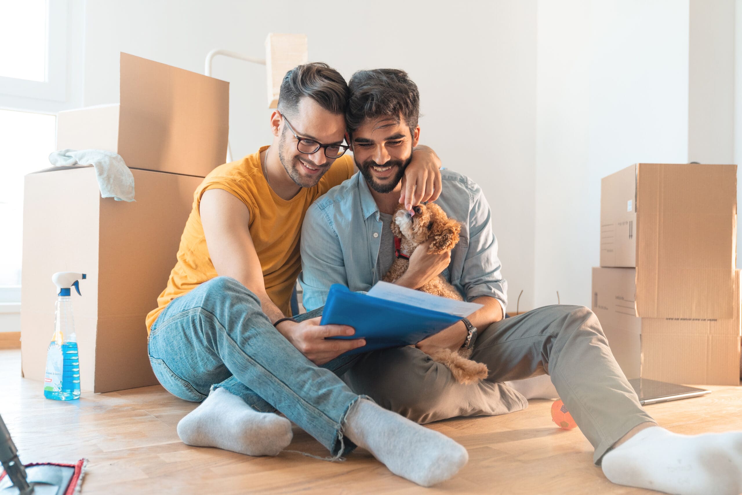 Happy gay couple smiling while cuddling dog and reading documents surrounded by cardboard boxes. Couple going through paperwork making financial plan together.